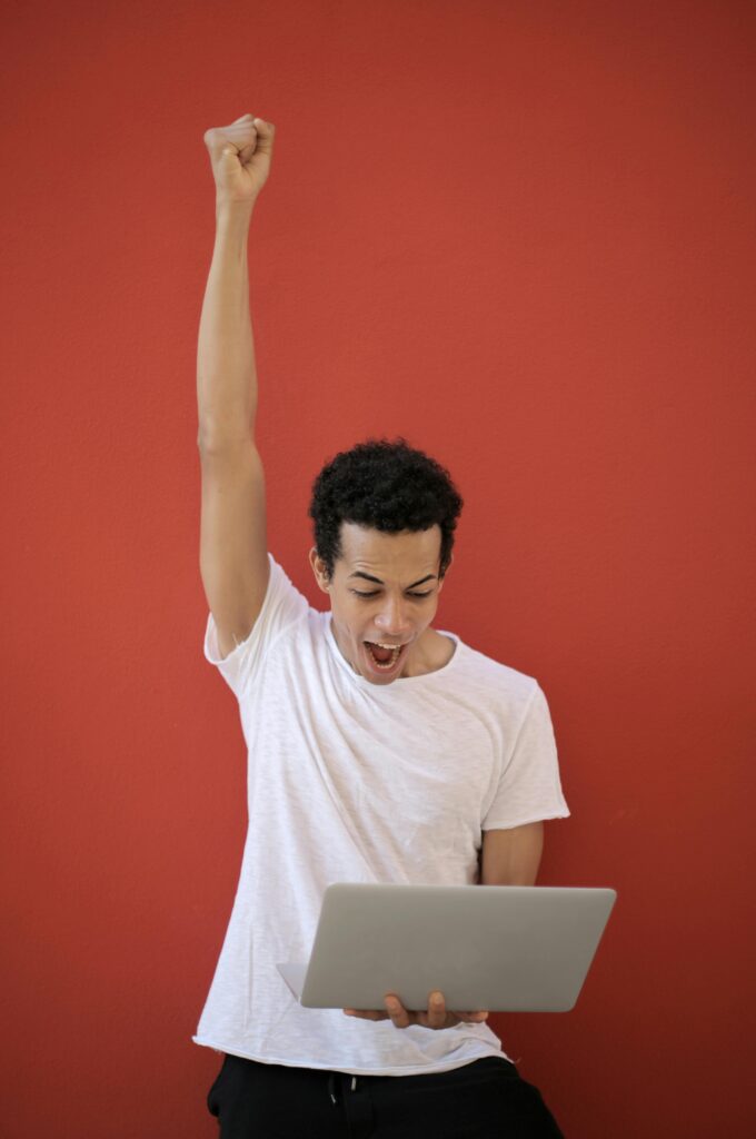 A young man in a white shirt celebrates success holding a laptop against a red background.